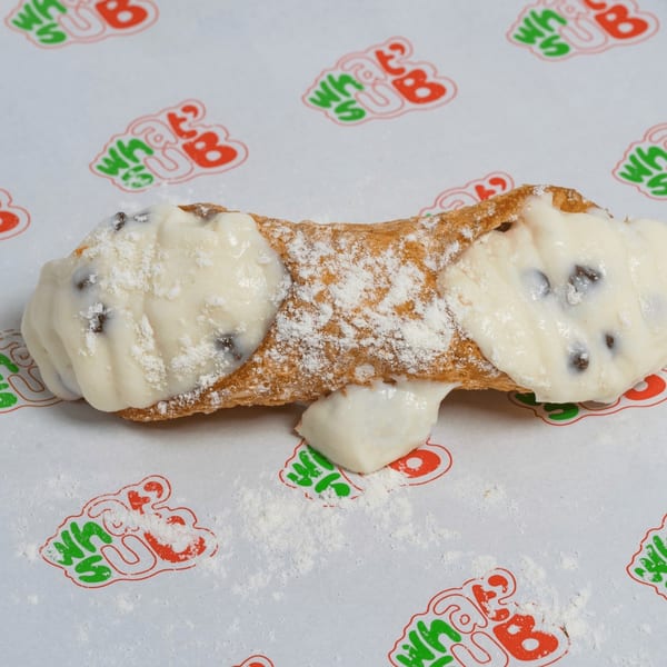 A cannoli filled with white cream and chocolate chips, dusted with powdered sugar, on a branded paper.