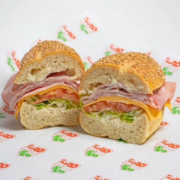 Two halves of a deli sandwich with ham, cheese, lettuce, and tomato on a sesame seed bun.
