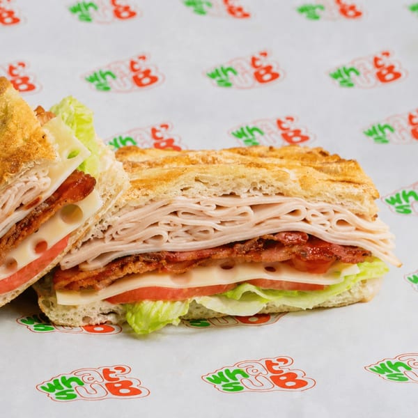 A turkey, bacon, and cheese sandwich with lettuce and tomato on toasted bread, on What Sub branded paper.