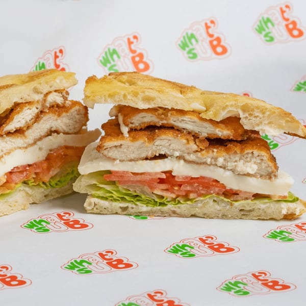 Two halves of a What's Sub chicken sandwich with lettuce, tomato, and cheese on branded paper.
