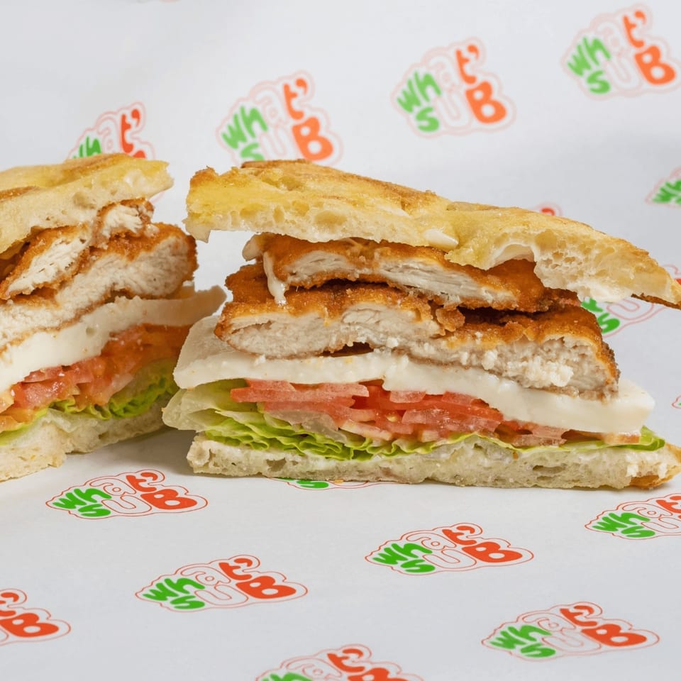 Two halves of a What's Sub chicken sandwich with lettuce, tomato, and cheese on branded paper.