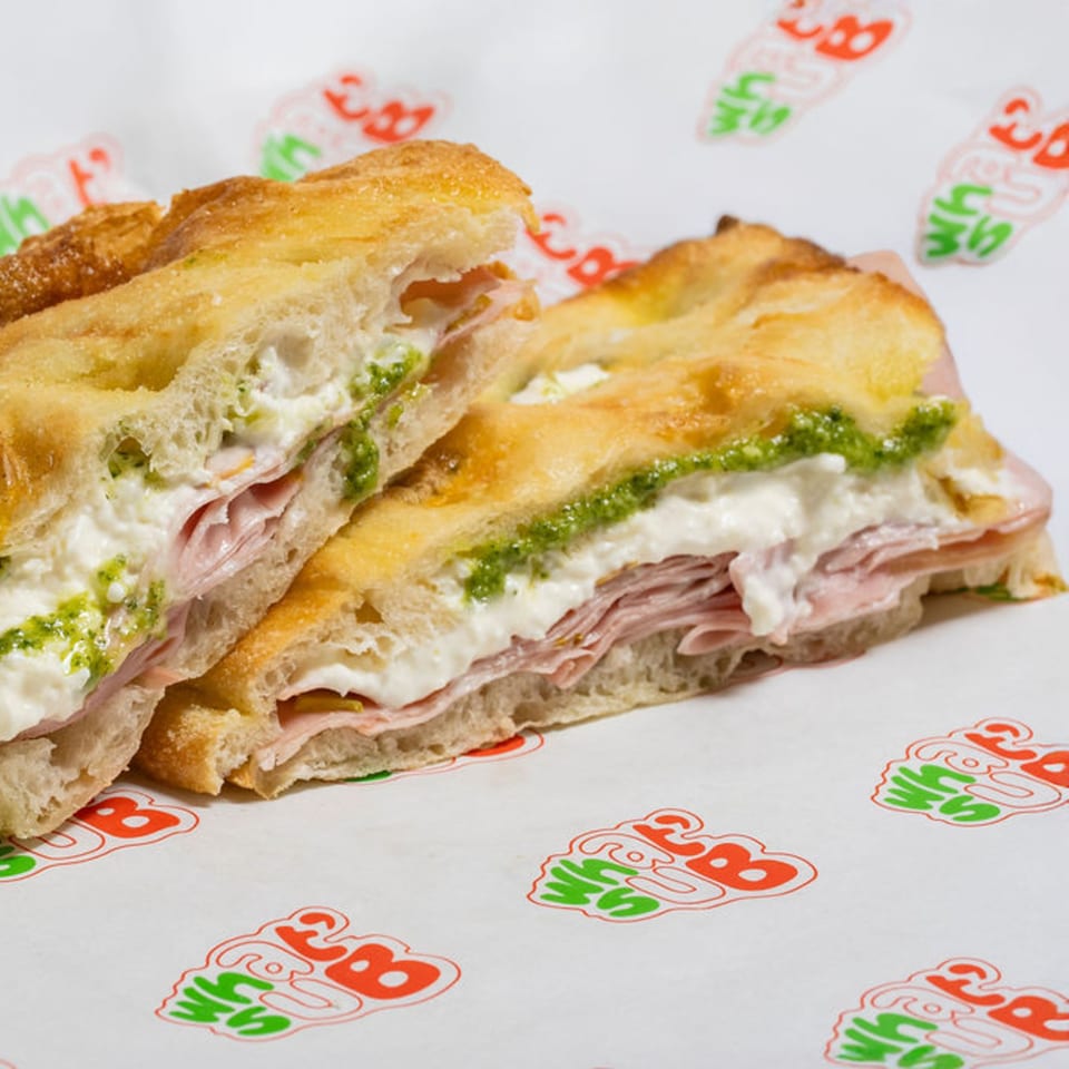 WhatSub Sub Shop's Italian focaccia sandwich with ham, creamy cheese, and pesto on branded paper.