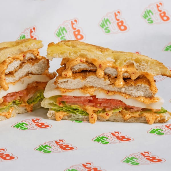 Two halves of a chicken sandwich with lettuce, tomato, cheese, and spicy sauce on What's Up branded paper.