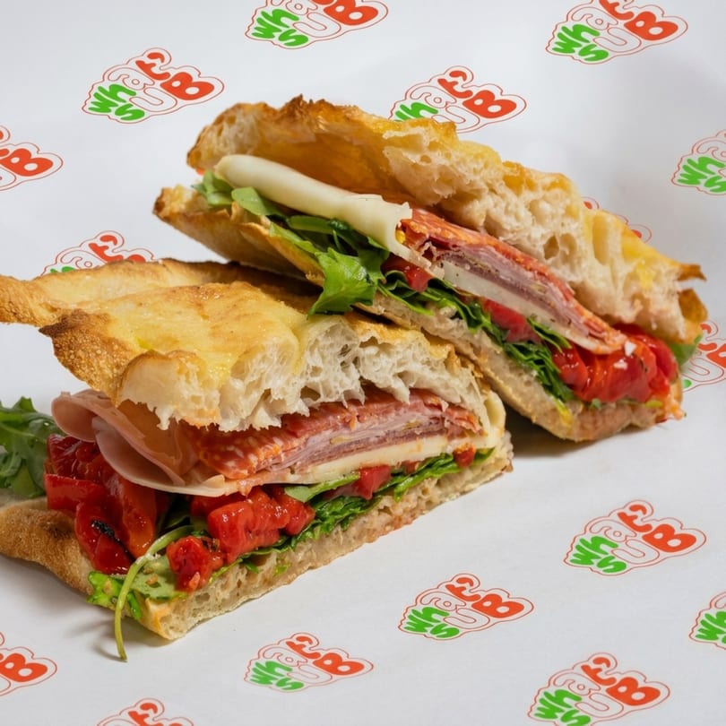 Two halves of a focaccia sandwich filled with deli meats, cheese, arugula, and roasted red peppers.