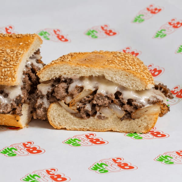 A sliced cheesesteak sandwich with melted cheese and onions on a sesame seed bun, on branded paper.