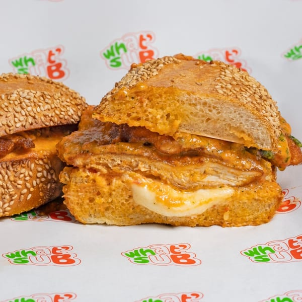 Two What's Up subs with sesame seed buns, chicken, melted cheese, and orange sauce on branded paper.