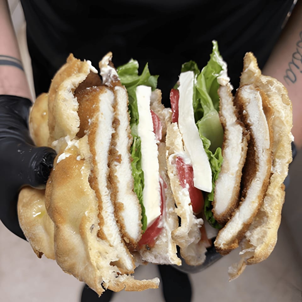 Hands in black gloves hold a large, sliced chicken cutlet sub with lettuce, tomatoes, and cheese.