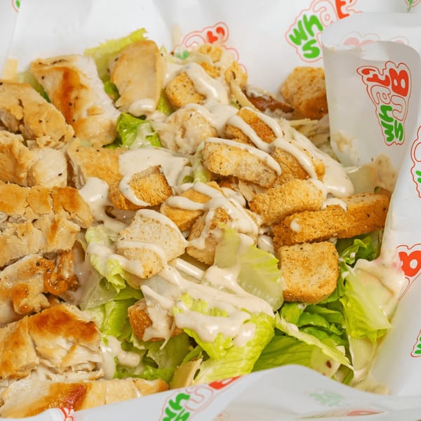 Caesar salad with grilled chicken, croutons, and dressing in a branded paper container.
