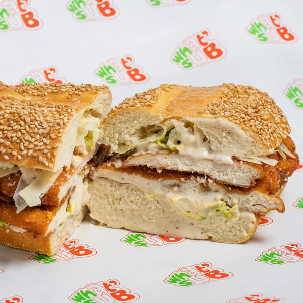 A chicken schnitzel sub, cut in half, with creamy sauce, lettuce, and cheese on a sesame seed bun.