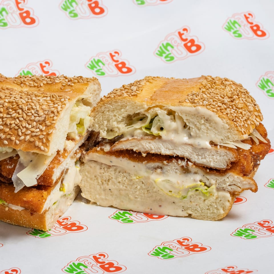 A chicken schnitzel sub, cut in half, with creamy sauce, lettuce, and cheese on a sesame seed bun.
